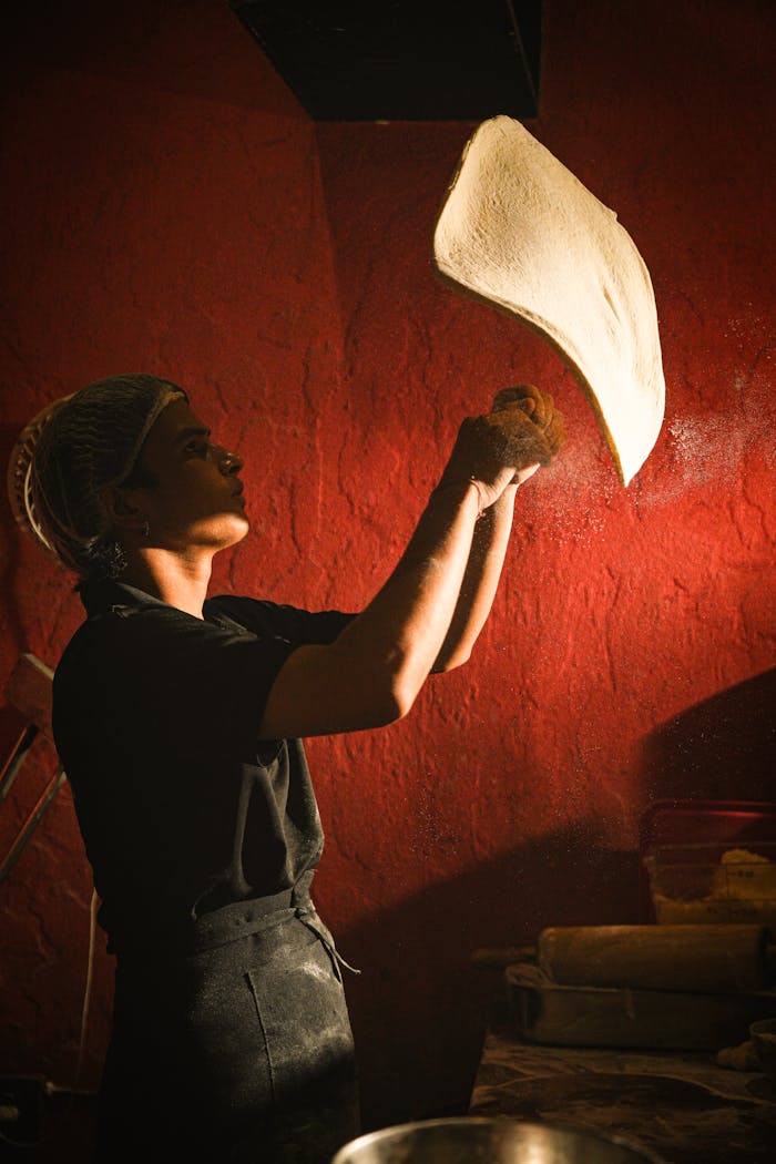 services-02 A chef skillfully tosses pizza dough in a warmly lit kitchen, showcasing traditional culinary artistry.